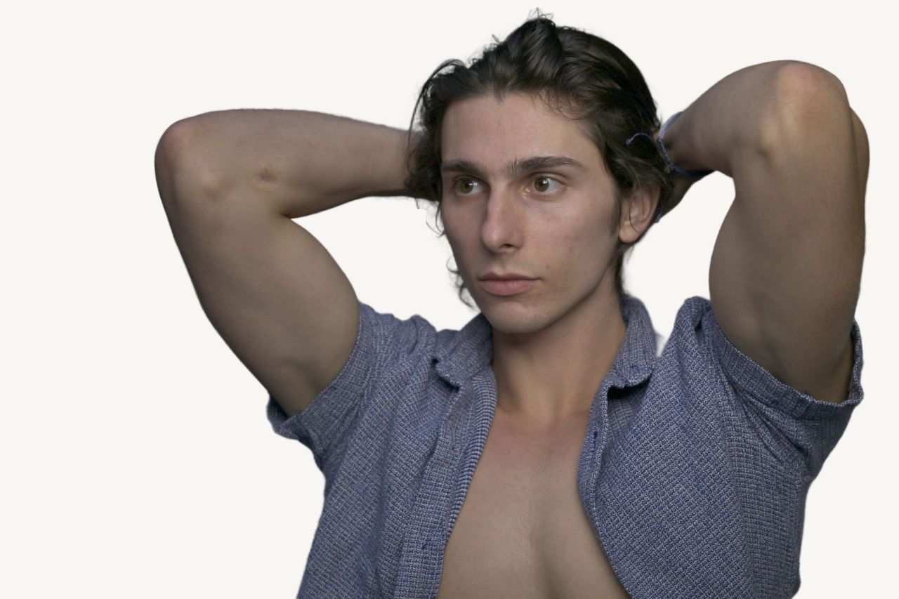 Tristan Colt, headshot 7: Tristan Colt with both hands behind his head, looking off to the side, open blue shirt showing an athletic build.