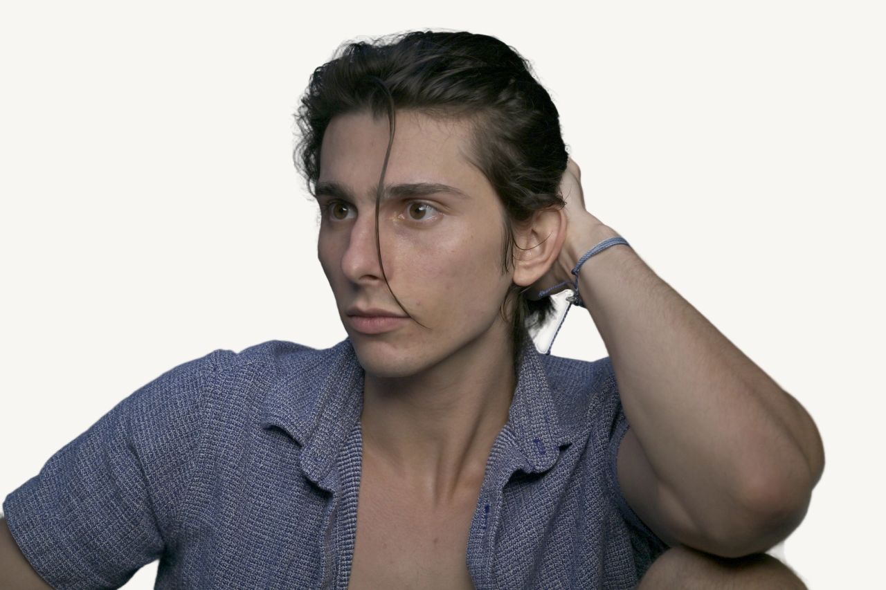 Tristan Colt, headshot 8: Tristan Colt with one hand in his hair, a strand falling across his face, glancing to the side, blue shirt.