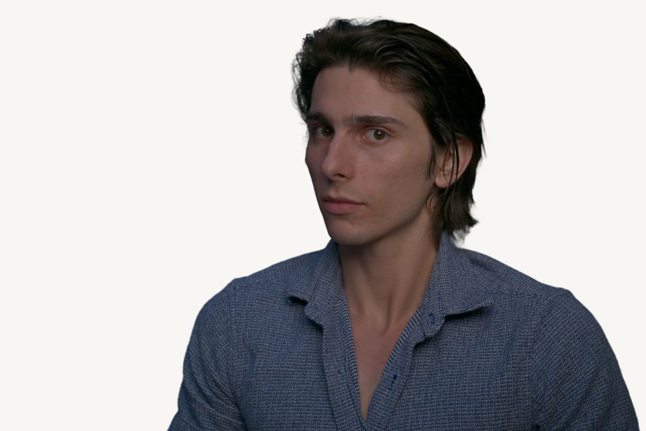 Tristan Colt, headshot 6: Tristan Colt turned slightly, looking past the camera with a pensive expression, open-collar blue shirt.