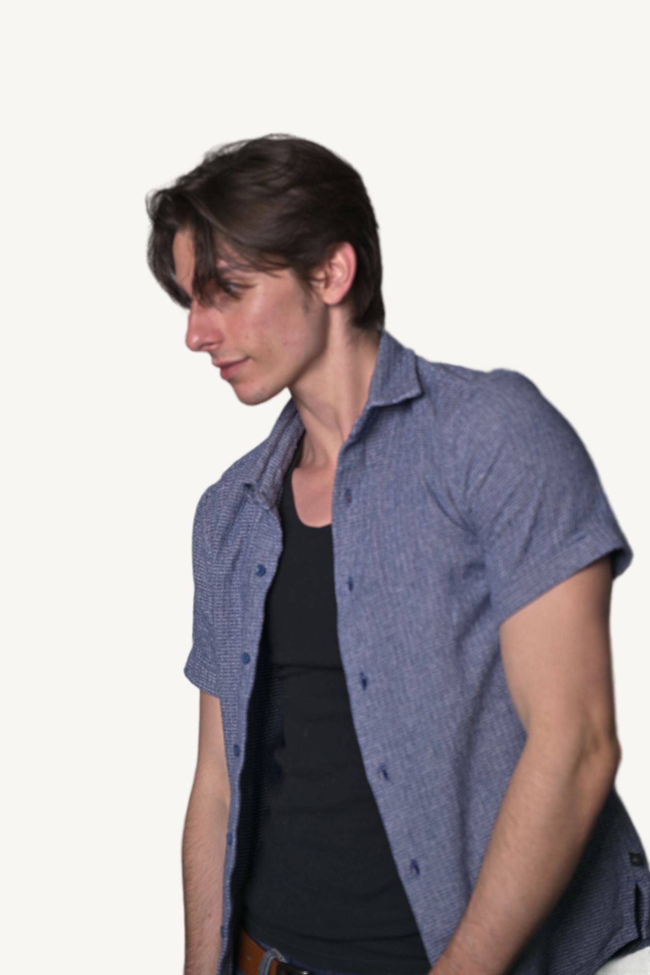 Tristan Colt, headshot 5: Tristan Colt standing, turned three-quarters and looking down, blue short-sleeve shirt open over a black tank top.